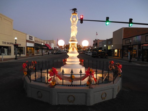 A decorative fountain with a statue at dusk, surrounded by shops and illuminated by streetlights and glowing orbs.