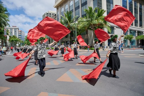 A parade scene with performers in black and white outfits waving large red flags amidst palm trees and buildings.