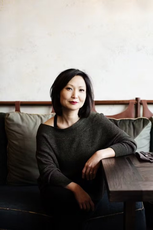 A woman with shoulder-length dark hair sits casually at a table, wearing a cozy sweater, with a relaxed expression.