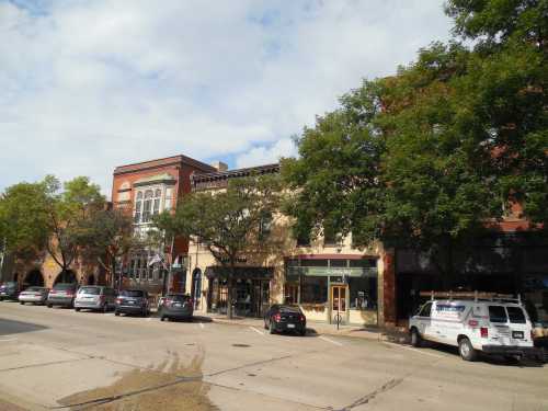 A street view of a small town with shops, trees, and parked cars under a partly cloudy sky.