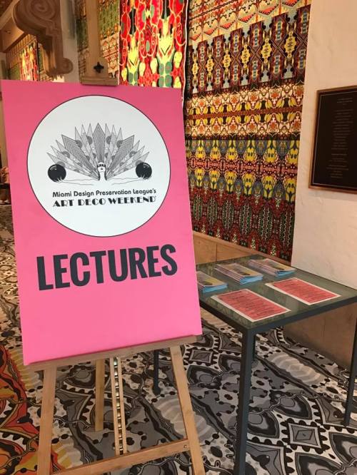 A pink sign for Miami Design Preservation League's Art Deco Weekend lectures, with colorful patterned fabric in the background.