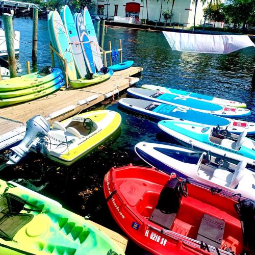 Colorful kayaks and paddleboards lined up on a dock by the water, with boats and a white sail in the background.