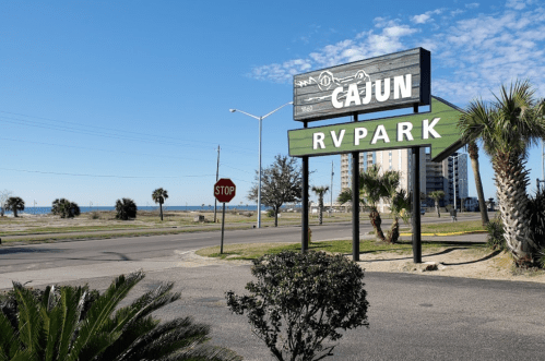 Sign for Cajun RV Park with palm trees and a view of the beach in the background under a clear blue sky.