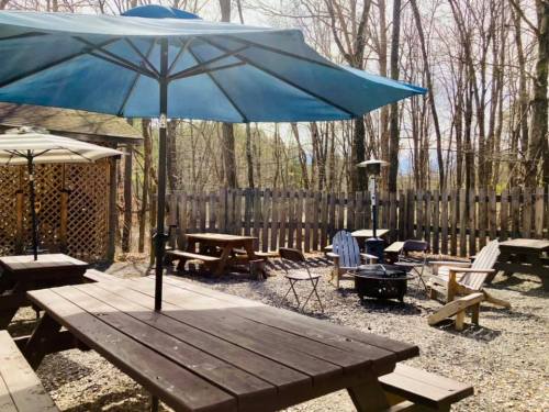 Outdoor seating area with picnic tables, umbrellas, and a fire pit, surrounded by trees and a wooden fence.