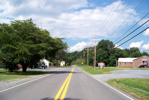 A rural road lined with trees and power lines, leading to distant hills under a partly cloudy sky.