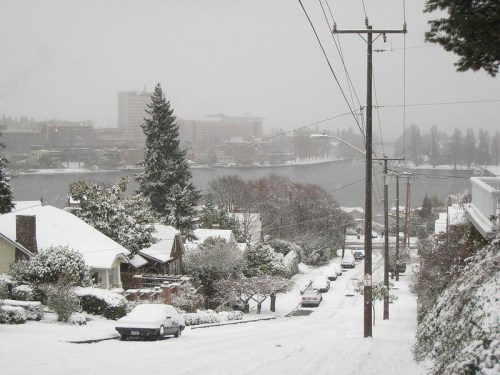 A snowy street scene with houses, trees, and power lines, overlooking a lake and distant buildings in a winter landscape.