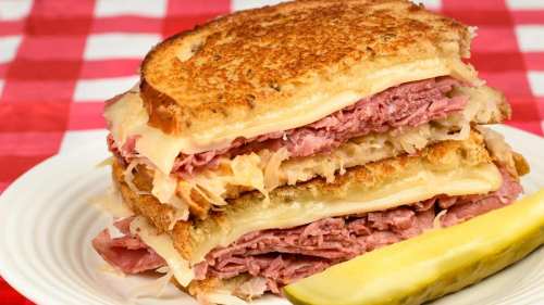 A stacked sandwich with layers of ham, cheese, and sauerkraut, served with a pickle on the side, on a white plate.