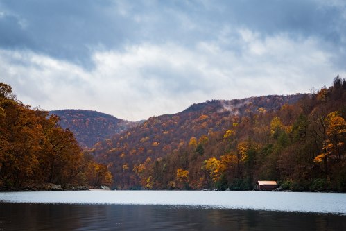 A serene lake surrounded by autumn trees, with misty mountains in the background under a cloudy sky.