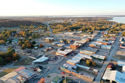 Aerial view of a small town with buildings, streets, and trees, surrounded by autumn foliage and a lake in the distance.