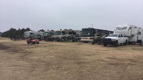 A row of parked RVs and trailers on a grassy area under a cloudy sky. A small vehicle is visible in the foreground.