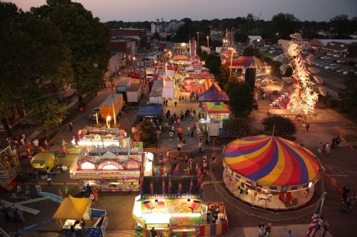 Aerial view of a vibrant carnival at dusk, featuring colorful tents, rides, and crowds enjoying the festivities.