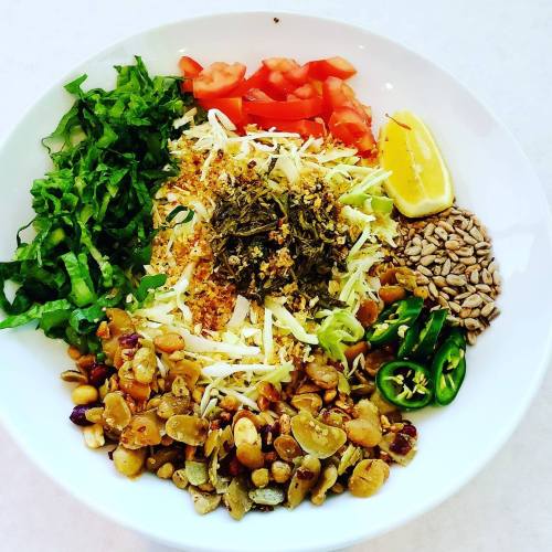 A colorful salad bowl with greens, tomatoes, cabbage, herbs, lemon, sunflower seeds, and sliced jalapeños.
