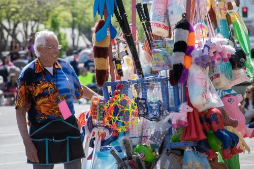 An elderly man stands beside a colorful booth filled with toys and cotton candy at a festive outdoor event.