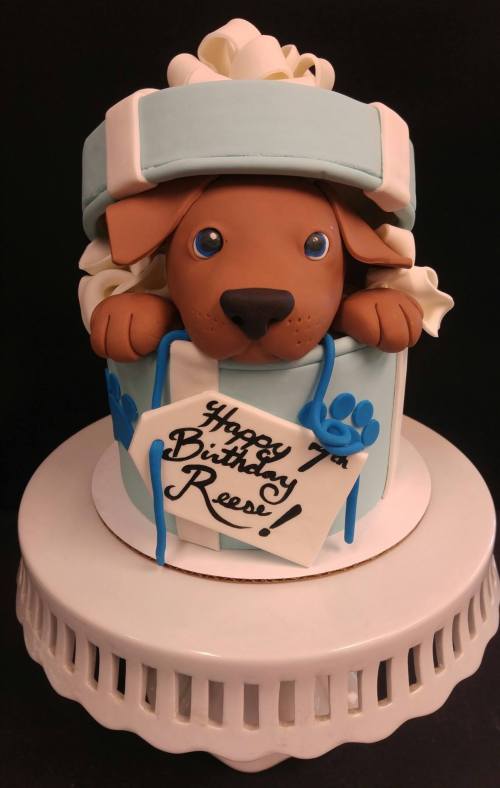 A cute cake featuring a puppy in a gift box, with a sign that says "Happy 7th Birthday Reese!" in blue icing.