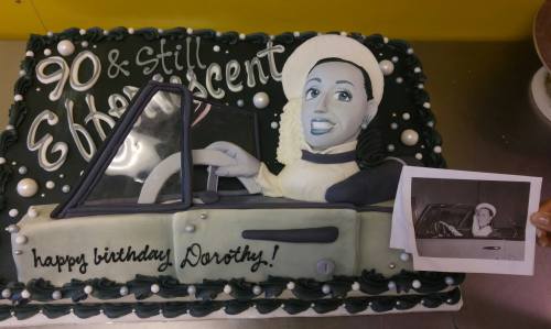 A birthday cake featuring a 3D figure of a woman in a car, celebrating Dorothy's 90th birthday with a photo beside it.