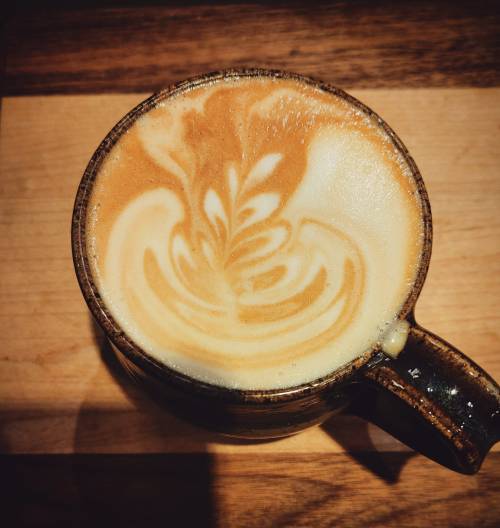 A close-up of a latte with intricate foam art in a dark ceramic mug on a wooden surface.