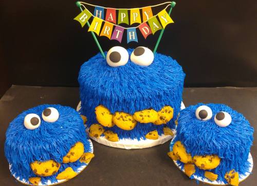 A colorful blue birthday cake decorated with cookie monster features and cookies, topped with a "Happy Birthday" banner.