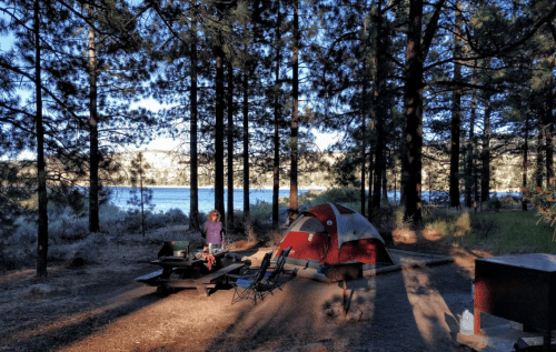 A campsite with a red tent, picnic table, and a person standing near a lake surrounded by tall trees.