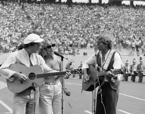 Three musicians perform on stage with guitars in front of a large crowd at an outdoor venue.