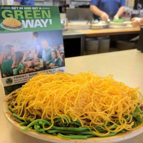A plate of green noodles topped with chili and a mound of shredded cheese, with a promotional sign in the background.