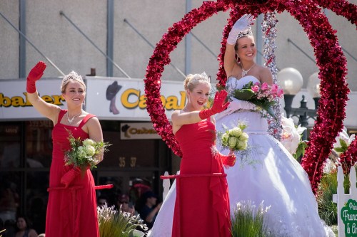 Three women in red dresses and gloves wave from a decorated parade float, with a festive backdrop.