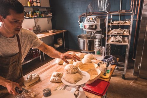 A baker shapes dough on a wooden table in a well-equipped kitchen, with various tools and ingredients around.