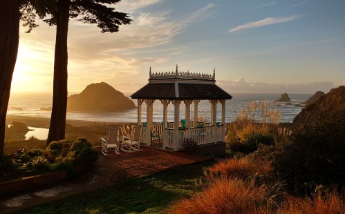 A gazebo overlooks a scenic beach at sunset, with waves crashing and distant cliffs in the background.