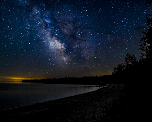 A starry night sky with the Milky Way over a calm lake and a dark shoreline.