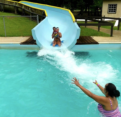 A child and adult slide down a water slide, splashing into a pool while another person reaches out from the water.