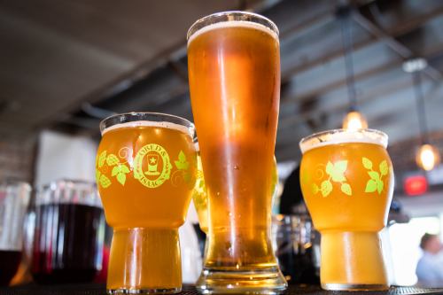 Three glasses of beer, two short and one tall, with a rustic bar background and warm lighting.