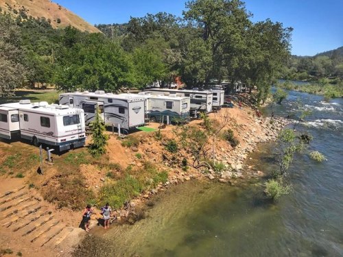 A scenic riverside campsite with RVs parked along the shore, surrounded by trees and a clear blue sky.