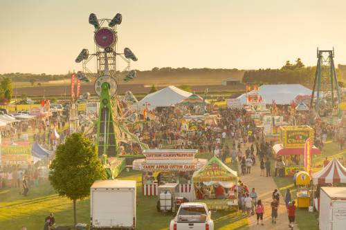 A bustling fairground at sunset, filled with people, colorful booths, and carnival rides.