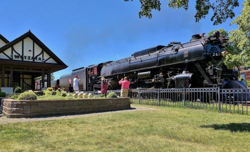 A vintage steam train at a station, with people taking photos and enjoying the sunny day. Green grass and trees surround the scene.