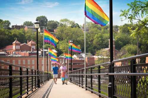 A couple walks hand-in-hand on a bridge adorned with rainbow flags, surrounded by greenery and buildings.