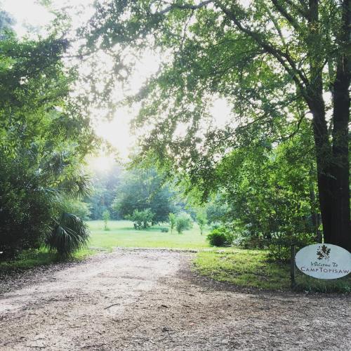 A gravel path leads to a sign reading "Welcome to Camp Topisaw," surrounded by lush greenery and soft morning light.