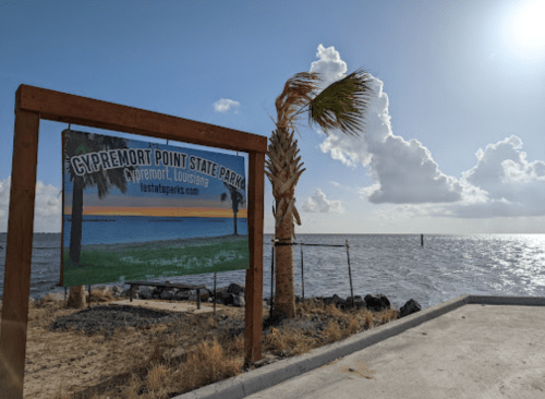 Sign for Cypremort Point State Park in Louisiana, with a view of the water and palm trees under a cloudy sky.
