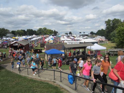 A bustling outdoor fair with crowds, tents, and vendors under a cloudy sky, viewed from a sloped pathway.