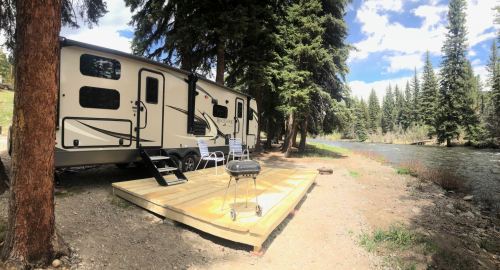 A camper trailer by a river, surrounded by tall trees, with a wooden deck and two chairs. A grill is nearby.