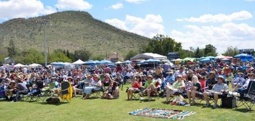 A large crowd sits on lawn chairs at an outdoor event, with tents and a mountain in the background under a blue sky.
