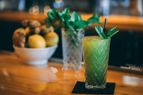 A refreshing green cocktail garnished with mint, placed on a wooden bar with lemons and ginger in the background.