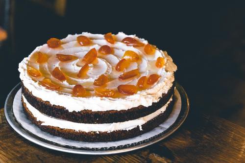 A layered cake with white frosting and topped with orange fruit pieces, displayed on a silver plate.