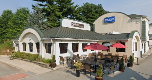 Exterior view of a restaurant with a green roof, outdoor seating, and red umbrellas, surrounded by trees.