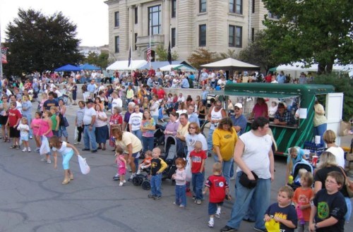 A large crowd gathers at an outdoor event with tents, food stalls, and families, including many children.