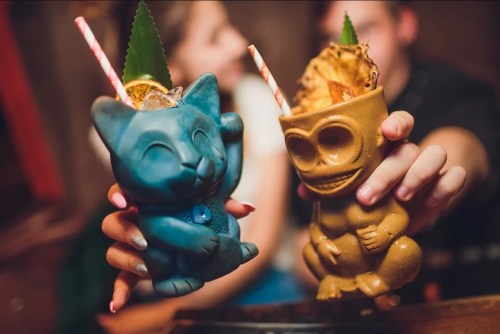 Two colorful tiki mugs shaped like animals, one blue and one gold, each with a straw and garnished with fruit.
