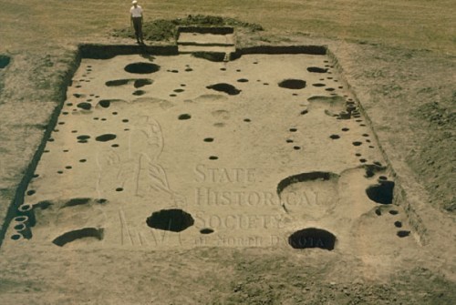 Aerial view of an archaeological dig site with numerous holes and features, with a person standing nearby.