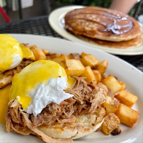 A plate of eggs Benedict with pulled pork and potatoes, with pancakes in the background.