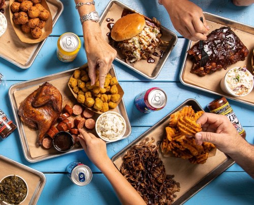 A vibrant spread of barbecue dishes, including chicken, ribs, sausage, and sides, with hands reaching for food.