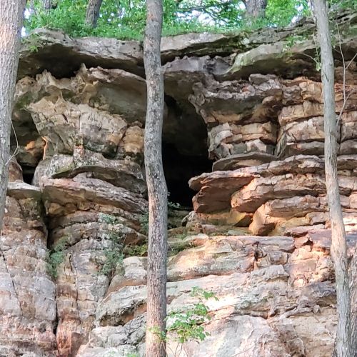 Rocky cliffside with layered stone formations and trees growing nearby. A small cave entrance is visible in the rock.