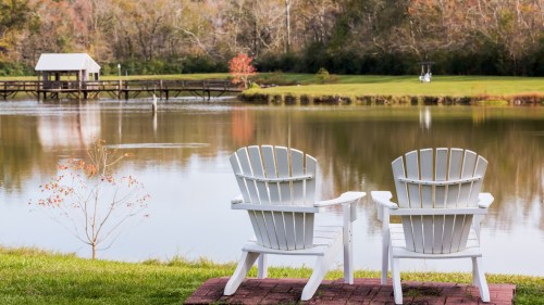 Two white Adirondack chairs facing a calm lake, surrounded by greenery and autumn trees in the background.