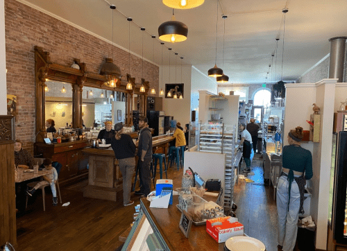 A cozy café interior with patrons at tables, a bar area, and a visible kitchen, featuring warm lighting and brick walls.